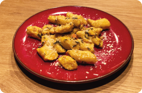 Teller-Kuerbis_Gnocchi Ein weißer Teller mit einem bunten Brotsalat aus gerösteten Brotstücken, gelber und grüner Zucchini, Kirschtomaten am Strunk und Auberginenstücken steht auf einem Holzbrett im Gras. Als Dekoration dienen große grüne Blätter
