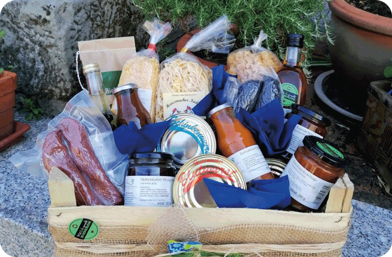 Drei Weinflaschen stehen auf einer kleinen Holzbank vor einer großen Feuerschale in Herzform. Hinter den Flammen ist ein gepflasterter Außenbereich zu sehen, umgeben von Holzstapeln und winterlicher Landschaft in der Dämmerung.